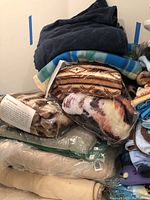 Stack of various fleece blankets in different colors and patterns, some wrapped in plastic bags.