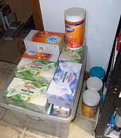 Photo showing four Clorox disinfecting wipes canisters along with six boxes of facial tissue including Puffs brand, arranged on and inside a clear plastic storage bin.