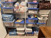Wide shot of three shelves with plastic storage containers, some blue boxes labeled, and a leopard print bag containing various crafting supplies and materials.