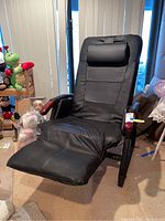 View of black leather-like padded massage recliner chair with wood armrests and cushioned headrest, positioned in a room with toys and furniture.