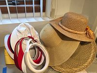 Group of women's hats and visors including fabric visors, white bucket-style hat, and several straw hats with decorative effects.