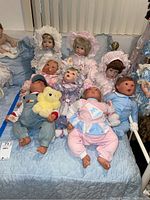 Full view of eight heirloom baby dolls on quilted blue surface showing varied outfits and accessories like plush toys.