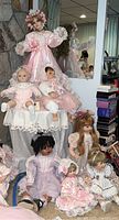 Full view of 7 heirloom dolls arranged on a lace covered table with background mirror and stone wall