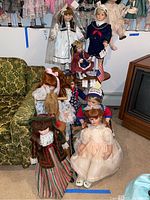 Group of heirloom dolls seated and standing near a green couch and television, showing variety of clothing and sizes