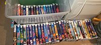 Top view showing two rows of VHS movies inside a plastic bin and arranged on the floor. Visible titles mostly Disney classics and children's movies.