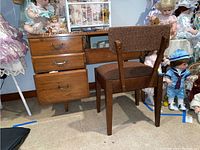 Front view of wooden cabinet with three drawers and brown upholstered chair in front, surrounded by dolls.