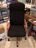 Front view of a black upholstered office chair with wooden armrests and base, placed in a room with various office supplies visible in the background.