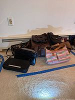 Multiple purses of different sizes, colors, and materials arranged on carpeted floor near a wall baseboard.
