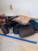 Wide view showing a pile on the floor of various purses in different shapes, colors, and sizes including black, brown, and beige tones.