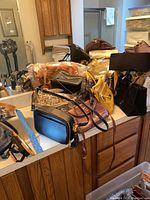 Multiple purses on a countertop showing various shapes and materials including a black rectangular purse with tassel, a metallic bronze purse, a yellow zippered purse, and others.