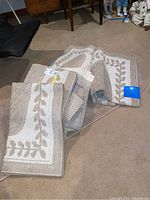 Two folded beige and cream bathroom rugs with leaf pattern on floor near furniture.