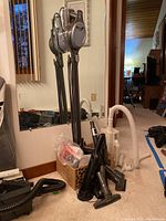 Two vacuums, Shark cordless stick vacuum mounted on a charging dock with accessories, and Oreck upright vacuum with hoses and attachments, stored upright near wall.