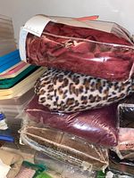 Stack of various plush throws and wraps including a burgundy package, leopard print throw, and a brown textured plush wrap.