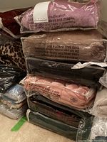 Stack of various plush wraps and throws in plastic packaging including pink, brown, black colors.
