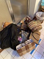 Photo showing assortment of totes and purses piled on a tiled floor including woven bags, a black drawstring bag, and a khaki nylon tote with brown trim.