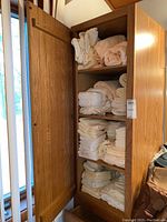 Wooden linen cabinet with four shelves filled with neatly stacked mostly white and some pale pink bath towels and washcloths.