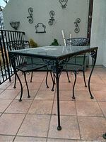 Angled front view showing metal scrollwork legs and clear glass top with chairs in background
