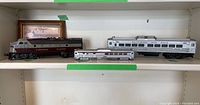 Three model train pieces and a framed photo of a train engine arranged on white shelves