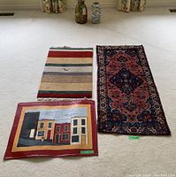 Three rugs laid on floor: wool pile runner with pattern, house design mat with red border, and colorful striped mat with fringes and small embroidered animal