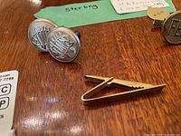 Pair of round sterling silver cufflinks with emblem design and plain sterling silver tie clip on wooden surface.