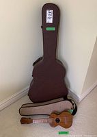 Hard shell guitar case standing upright with padded ukulele case and ukulele at base