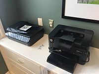 Both printers shown side-by-side on a white cabinet surface against a green wall with a framed picture.