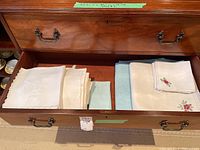 Top view of open drawer showing neatly stacked embroidered and lace table linens and napkins in white, cream, and blue colors.