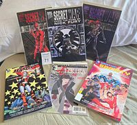 Six comic books displayed on couch, covers clearly visible including Marvel Secret War and DC Harley Quinn and Earth 2 Society.
