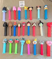 Top-down photo showing 26 assorted PEZ dispensers with heads including Star Wars, Disney characters, and others arranged in rows on a beige surface.
