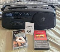 Front view of black Lenoxx Sound cassette recorder player with control buttons, speaker, AM/FM tuner, and two cassette tapes displayed in front of it on a fabric surface.