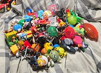 Pile of assorted toys including character figurines, vehicles, and novelty toys on a checkered fabric surface.