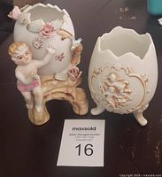Photo showing two ceramic cracked egg-shaped items. Left piece is a planter with a cherub holding the egg decorated with pink flowers and a bluebird. The right piece is a three-footed cracked egg vase with cherub relief design.