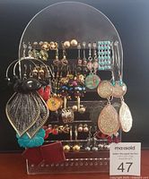 Photo of assorted costume earrings displayed on a clear acrylic earring holder, showing a variety of styles and colors including large leaf shapes, colorful beads, metallic tones, and glitter elements.