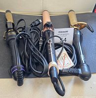 Three assorted curling irons with power cords, different barrel sizes and styles, placed side by side on a surface.