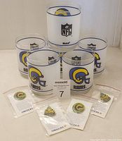 Lot of six clear drinking glasses featuring blue rim and St. Louis Rams helmet logo and NFL branding, arranged in a pyramid with four collectible pins in original Unocal packaging in front.