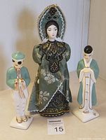 Front view of Japanese female porcelain figure in detailed green and black kimono and two male Japanese porcelain figures, one playing flute and one standing beside her.