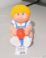 Front view of the piggy bank doll, showing facial features, yellow hair, and holding an orange ball.