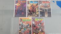 Five Spawn comic books laid out flat on a surface, bagged and boarded, showing colorful covers with Spawn artwork.