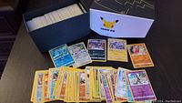 Photo showing a black trainer box filled with sleeved Pokemon cards, several cards displayed in front including Water, Psychic, Fire, Electric types and trainers.