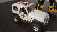 White Jeep Wrangler medical vehicle with red cross decals on doors and roof, metal body with detailed wheels and front grille shown on a dark flat surface.