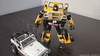 Yellow Transformers Jeep Wrangler robot figure standing upright next to white Jeep Wrangler vehicle toy on a black table surface.
