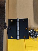 Single square matte black door hinge taken out of packaging showing flat hinge with four screw holes per leaf and central pin.