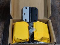 Door hinge resting on cardboard box with two yellow paper packages beneath.