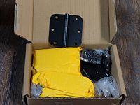 Box containing yellow wrapped and clear plastic wrapped matte black rounded door hinges, with one hinge visible outside of wrapping to show details.