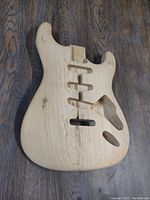 Top view of unfinished wooden Stratocaster guitar body on dark wood floor showing front routed cavities.