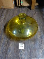 Top-down and angled side views of a large bowl-shaped vase made of amber yellow-green glass, showing its size and shape against a wood floor background.