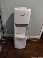 Front view of the white Primo water dispenser standing against a light-colored wall on dark wood floor. Shows controls and dual water spouts at the top and lower panel storage compartment.