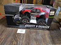 Front view of the Gravity V-Thunder RC truck in original box on wooden floor with lot number card visible.