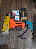 Photo showing all items in the lot on a wooden floor: Arrow electric stapler with power cord, Stanley glue sticks in packaging, Avery tagging tool, DeWalt tool pouch, and box of Arrow staples.