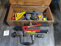 Wooden toolbox open showing various hand tools including bolt cutters, hand drills, hammer, and other tools.